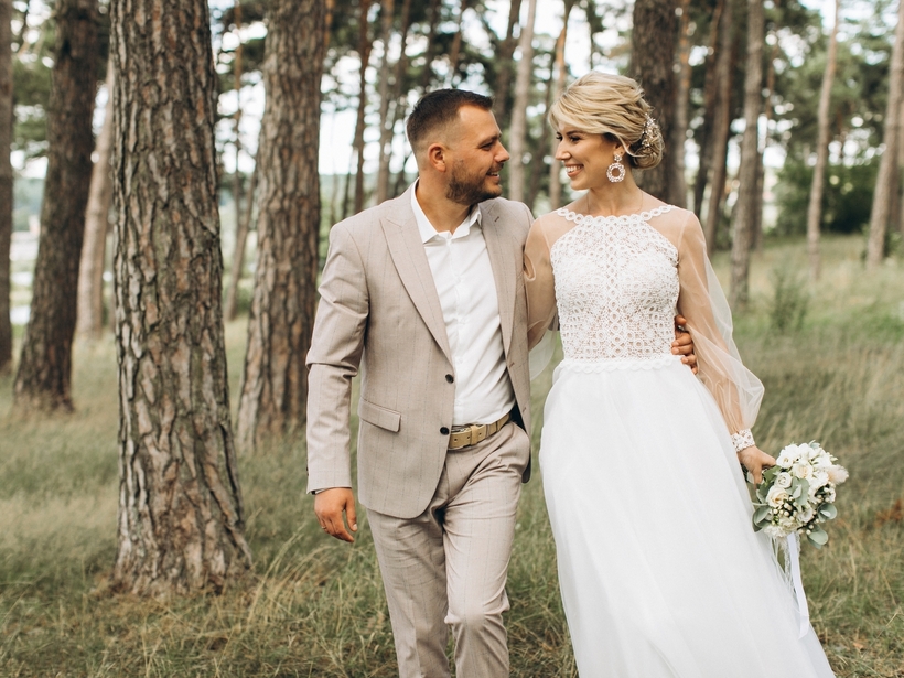 Newlyweds walking through pine woods after an outdoor wedding, bride in lace gown holding bouquet, groom in light beige suit