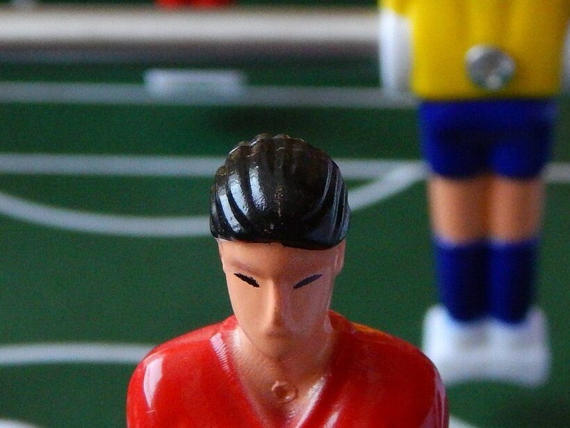 Nahaufnahme eines Tischkickers: roter Spieler vorne, verschwommene gelb-blaue Gegner auf dem grünen Spielfeld.