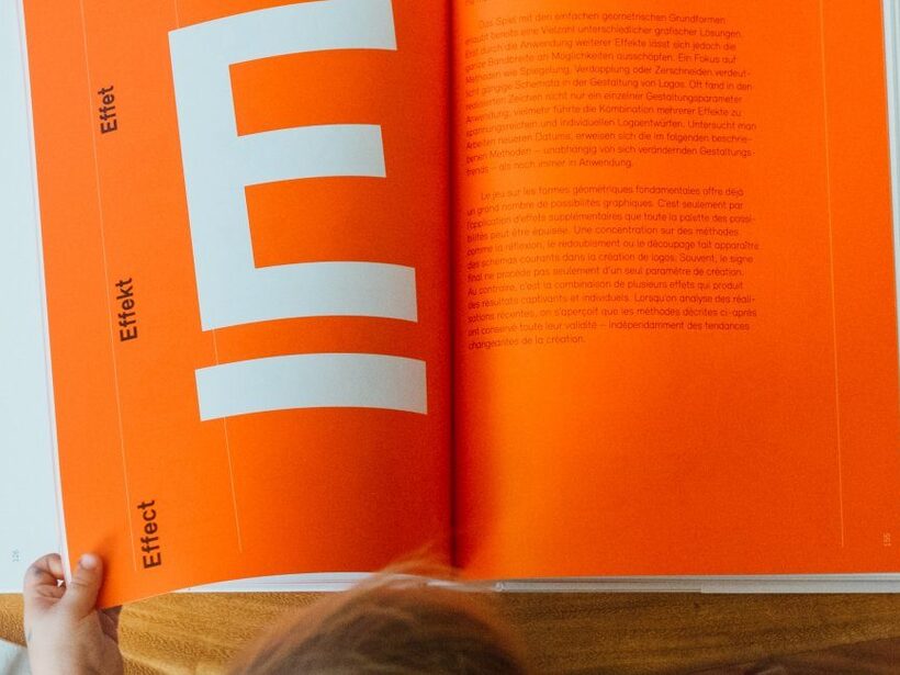 Kinderhand blättert in einem großen Designbuch mit leuchtend orangefarbenen Seiten auf einem Holztisch