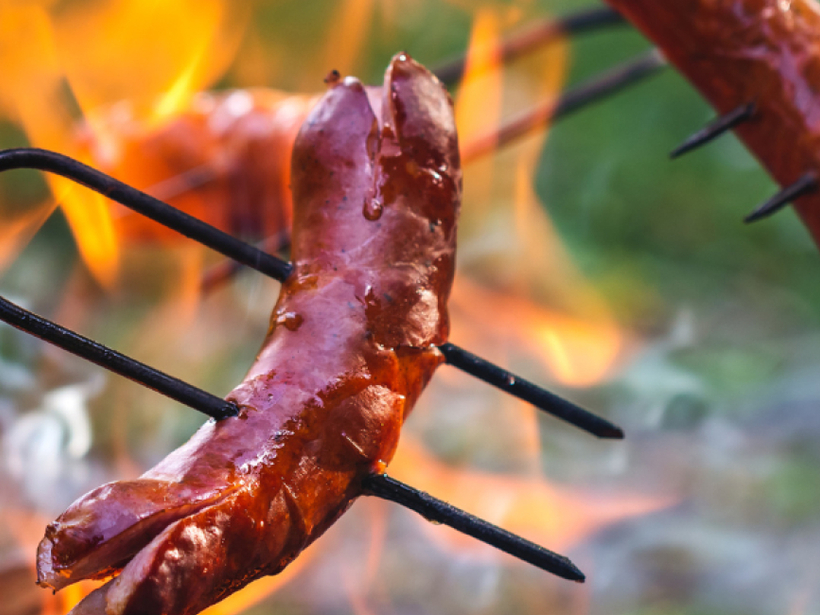 Saftige Würste auf Spießen über offenem Lagerfeuer, glänzende Haut und rauchiger Grillgenuss