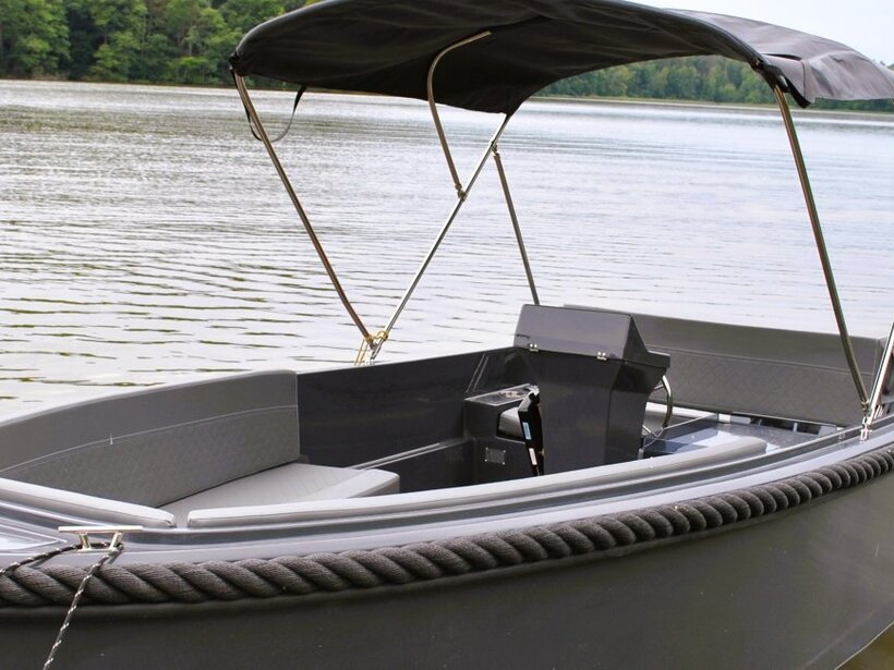 Kleines Freizeitboot mit gepolsterten Sitzen, Sonnendach und geflochtenem Fender auf ruhigem See vor bewaldetem Ufer