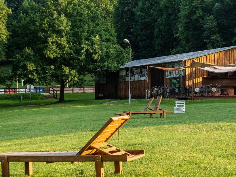 Sonnige Wiese mit Holzliegen und schattiger, rustikaler Terrassenbar am Waldrand für entspanntes Draußensitzen.