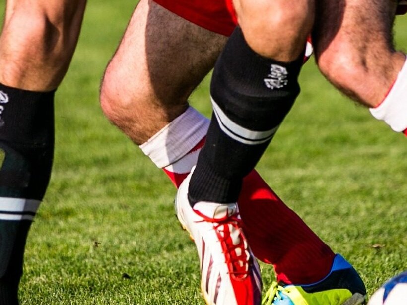 Beine zweier Fußballspieler in roten und schwarzen Stutzen, bunten Schuhen und Schienbeinschonern kämpfen um den Ball auf grünem Rasen