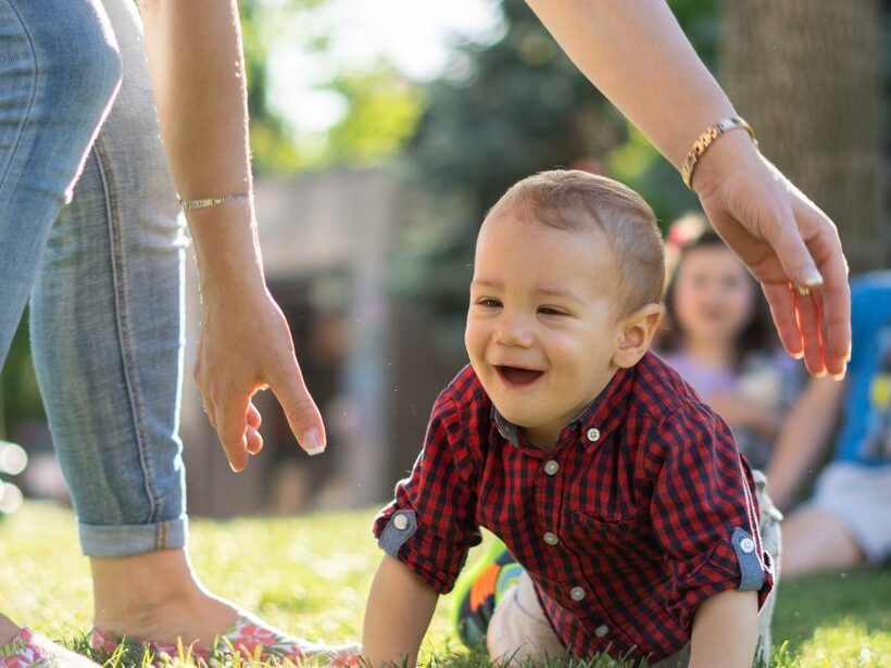 Lachendes Kleinkind krabbelt über sonnigen Hotelrasen, Eltern führen mit Händen, ältere Kinder entspannen im Hintergrund.