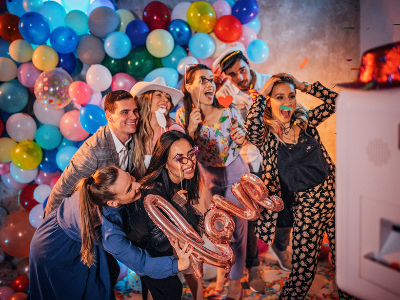 Lively hotel party in a decorated event space with balloon wall, confetti, photo booth and friends posing with props