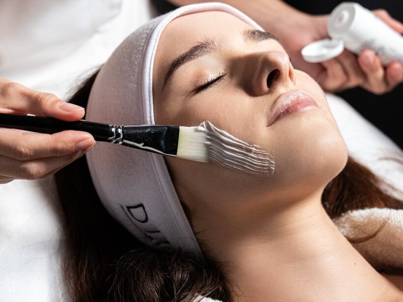 Gast liegt entspannt im Spa, Augen geschlossen, Kosmetikerin trägt mit Pinsel weiße Gesichtsmaske aus Tube auf; ruhige Behandlungsatmosphäre.