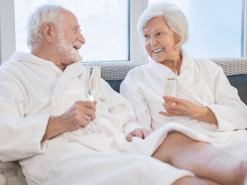 Älteres Paar in weißen Bademänteln lächelt auf einem Lounge-Sofa und stößt mit Champagner im hellen Spa-Entspannungsbereich an.