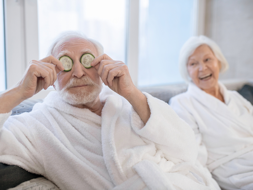 Älteres Paar in weichen weißen Bademänteln in der Spa-Lounge; Mann hält spielerisch Gurkenscheiben vor die Augen, Frau lacht.