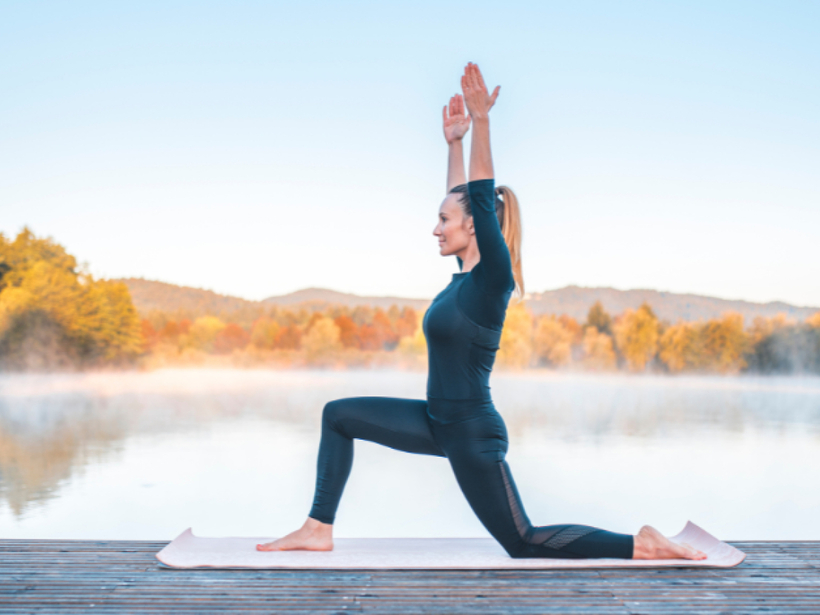 Frau macht morgendliches Yoga am See auf Holzsteg – ruhige Spa‑Aktivität mit Nebel über dem Wasser und herbstlichen Bäumen.