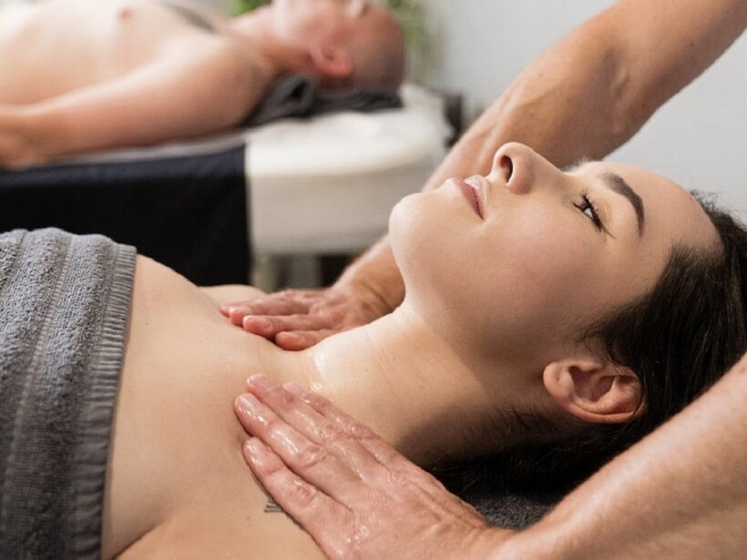 Entspannende Paarmassage im Spa, Frau liegt auf der Behandlungsliege, Therapeut massiert Nacken und Dekolleté, graues Handtuch