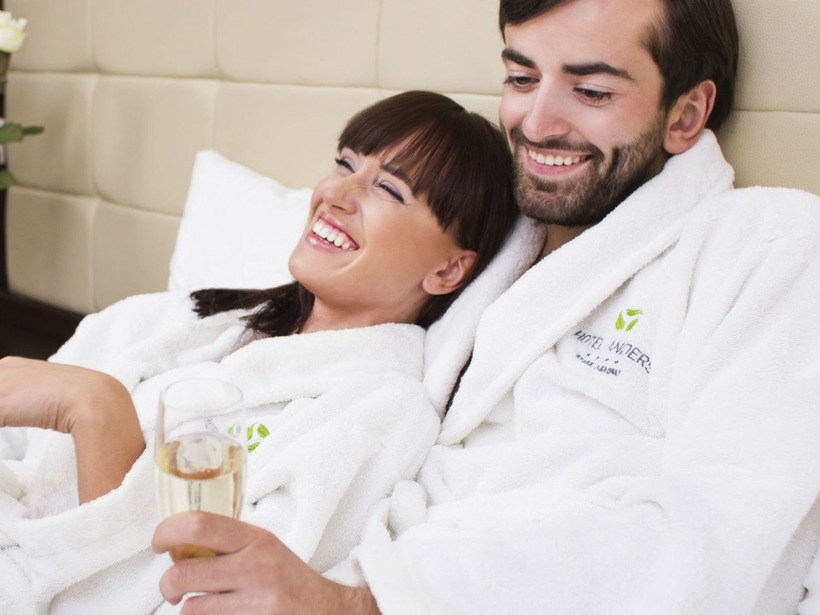 Paar in weißen Hotelbademänteln auf dem Bett, stößt mit Champagner an, frische Rosen am Nachttisch, entspannter romantischer Aufenthalt.