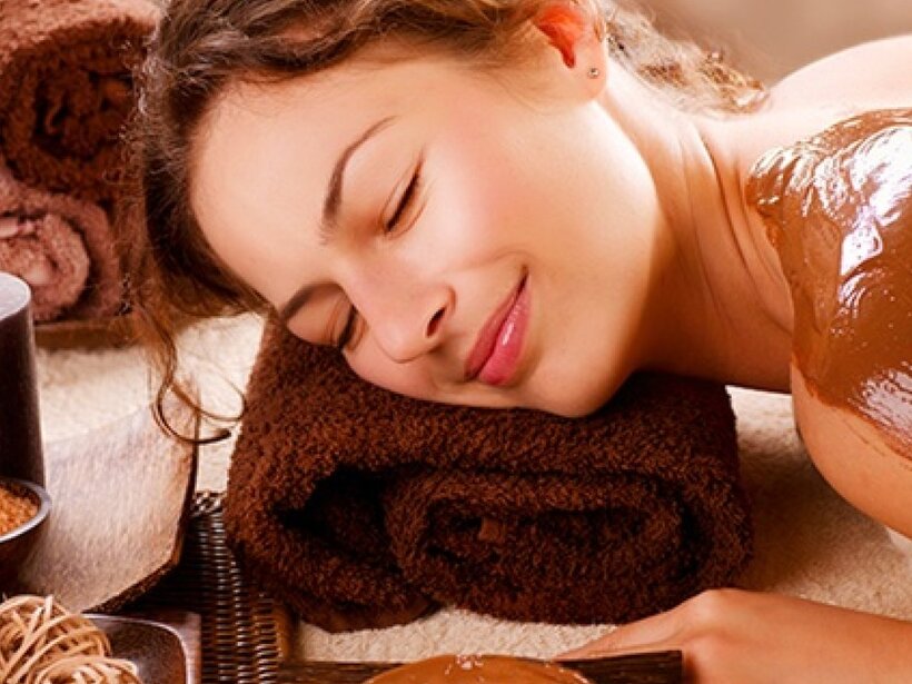Frau entspannt bei einer Schokoladen-Körperpackung auf dem Spa-Tisch, braune Handtücher, brennende Kerze und Badesalz.