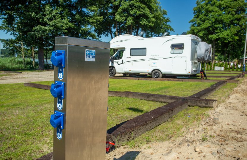 Stellplatz mit Edelstahl-Elektroanschluss und blauen Steckdosen, weißes Wohnmobil geparkt auf grasigem, baumbestandenen Platz.