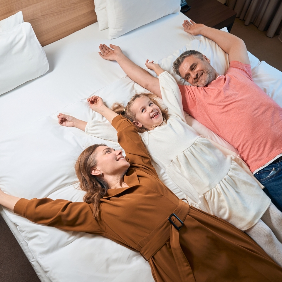 Eltern und Kind lächelnd auf einem King-Hotelbett mit weißer Bettwäsche, Holz-Kopfteil und Nachttisch ausgestreckt