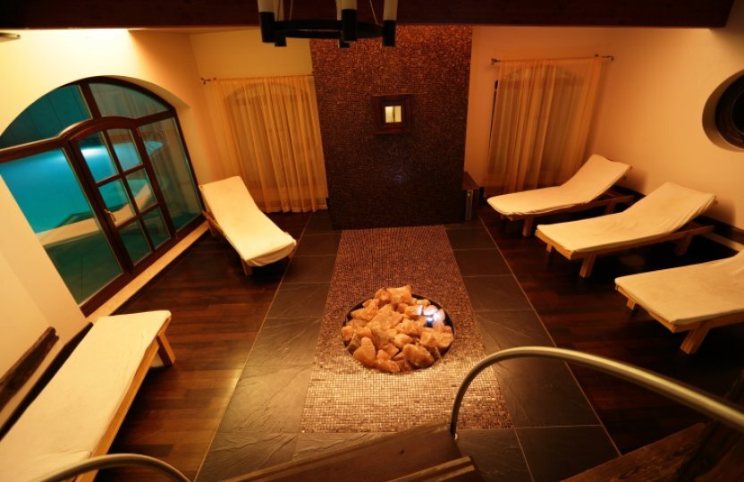 Gemütlicher Ruheraum im Spa mit Salzstein-Feuerstelle, gepolsterten Liegen und rundem Fenster zum Hallenbad.