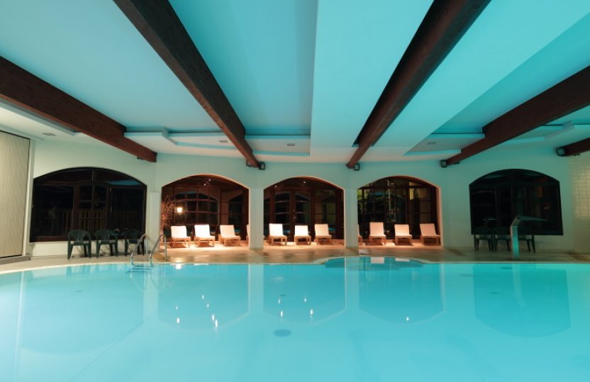 Hallenbad im Hotel mit türkisblauem Wasser, sichtbaren Holzträgern, rundbögigen Fenstern und Liegestühlen.
