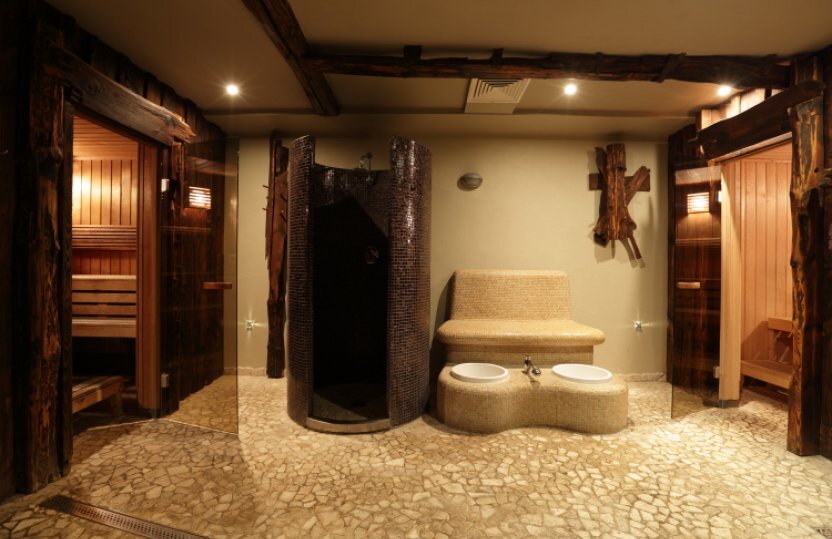 Spa-Bereich mit zwei Holzsaunen, zentraler Mosaikdusche, geschwungener Bank mit zwei Fußbecken und warmem Licht.