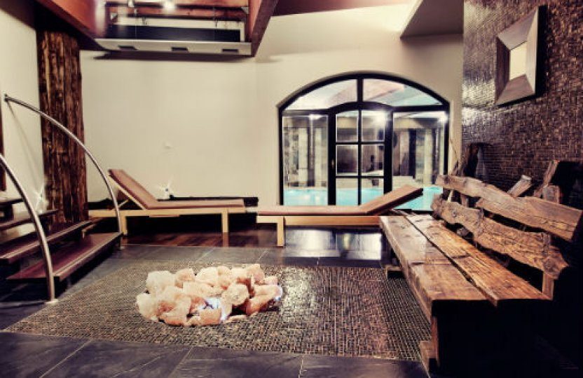 Spa-Lounge mit Holzbank und Liegen, Salzstein-Dekor auf Mosaikboden und bogenförmigen Glastüren zum Hallenbad.