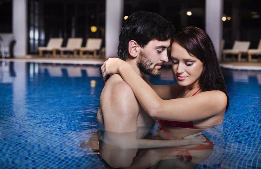 Paar umarmt sich im ruhigen Indoor-Hotelpool bei Nacht, Liegestühle und sanfte Beleuchtung im Hintergrund.