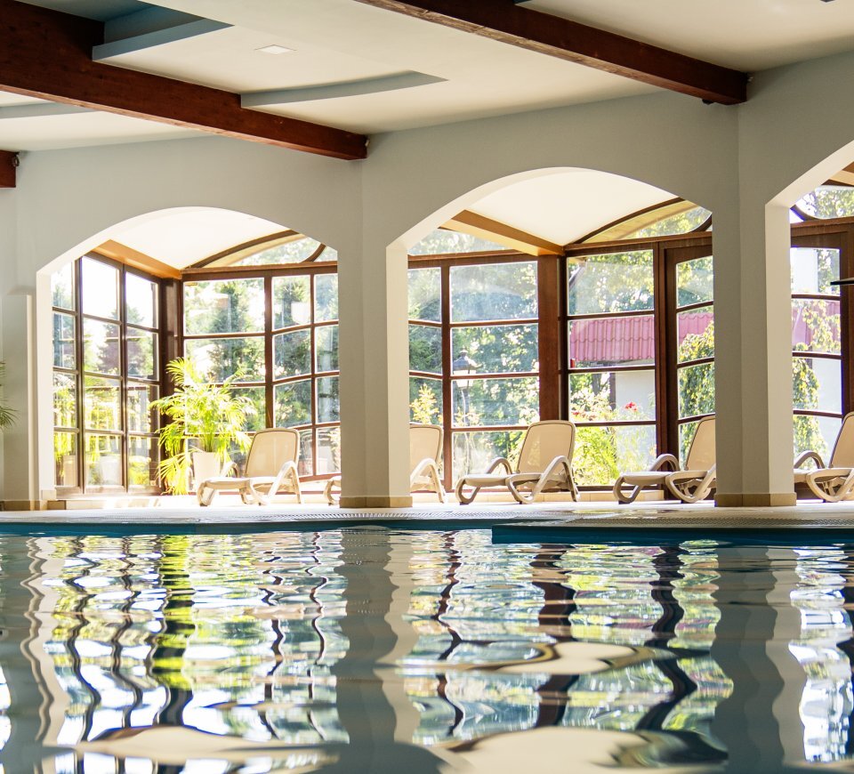 Überdachtes Hotel-Schwimmbad mit bogenförmigen Panoramafenstern, sonnigen Liegen, grünen Topfpflanzen und sichtbaren Holzdeckenbalken