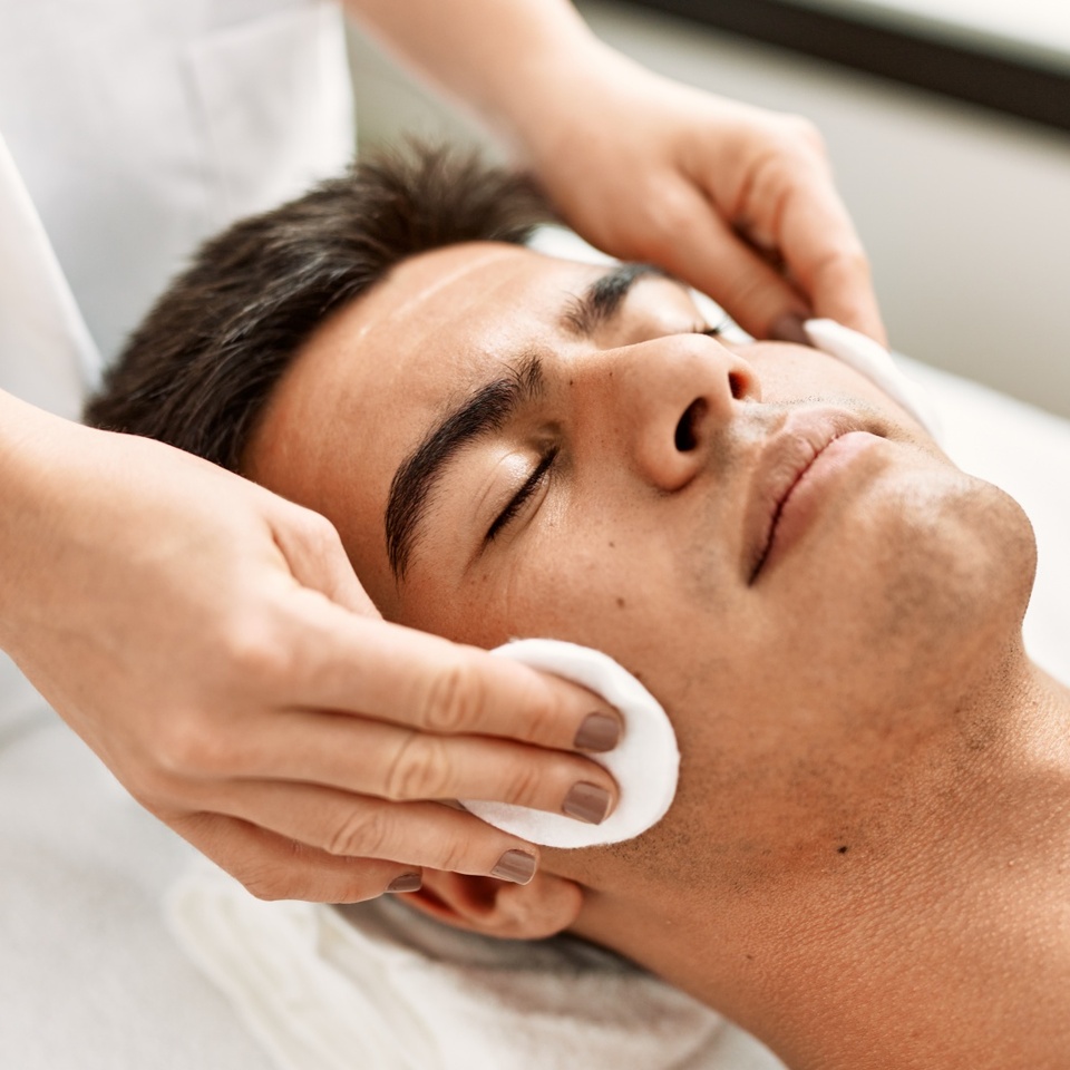 Entspannter Mann auf Spa-Liege, Kosmetikerin reinigt sein Gesicht mit Wattepads