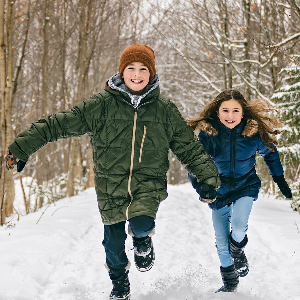 Two smiling children in winter coats running on a snow-covered forest trail, wearing boots, hats and gloves.