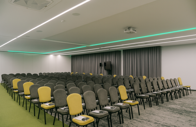 Nowoczesna sala konferencyjna w hotelu z rzędami szarych i żółtych krzeseł, projektorem sufitowym i zielonym oświetleniem.