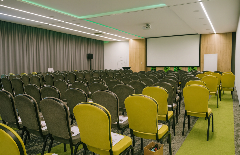 Hotel conference room with rows of gray and lime chairs, projection screen, podium, LED ceiling lighting and green aisle carpet
