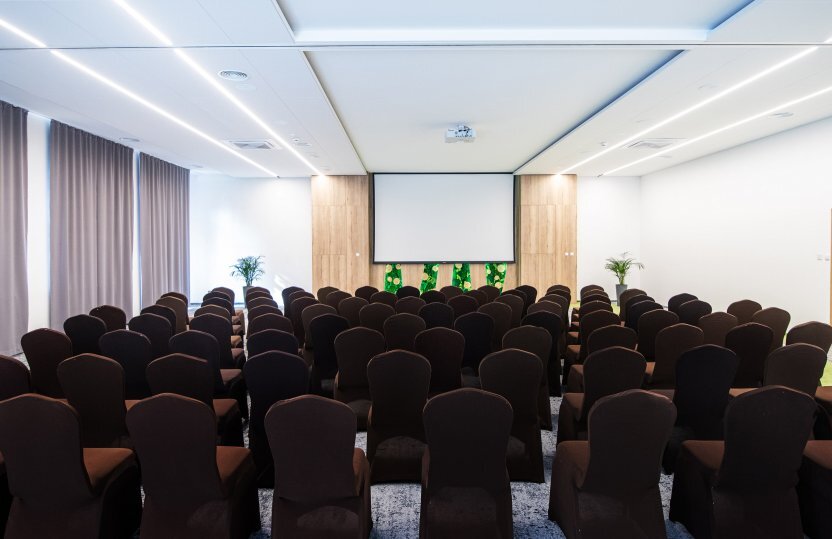 Nowoczesna sala konferencyjna hotelu z rzędami obitych krzeseł, ekranem projekcyjnym, drewnianą ścianą i roślinami