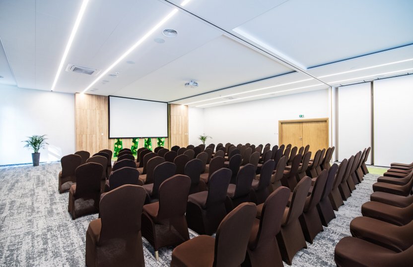 Nowoczesna sala konferencyjna w hotelu z rzędami brązowych krzeseł skierowanych na duży ekran projekcyjny, wykładzina i delikatne oświetlenie.