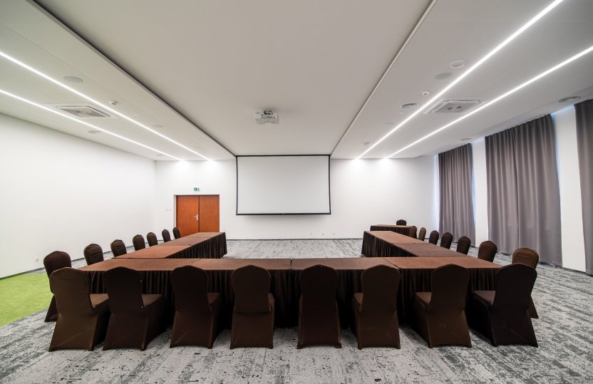 Spacious hotel conference room with U-shaped tables draped in brown, projector screen, ceiling lights and tall curtains