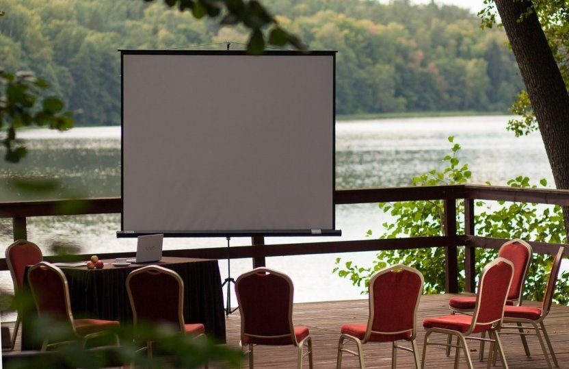 Lakeside wooden terrace set for a meeting with projection screen, red chairs, table with laptop and apples, peaceful forest and water view