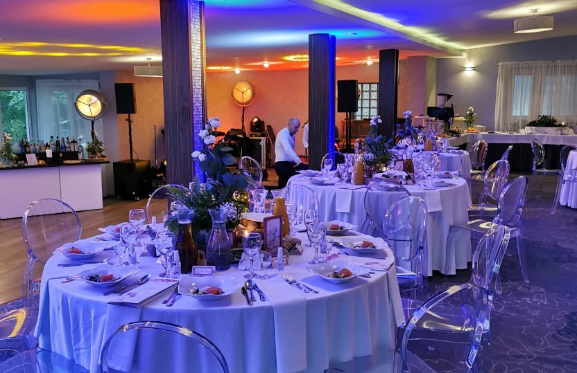 Banquet room with round tables in white linen, floral centerpieces, clear chairs, colorful uplighting and staff preparing service.