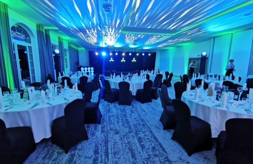 Hotelballsaal fürs Bankett mit runden Tischen, Tischdecken, schwarzen Stuhlhussen und Gedeck; Personal bereitet Bühne mit blau-grünem Licht vor.