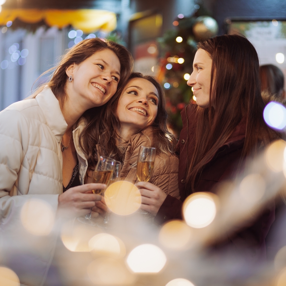 Drei Frauen in Wintermänteln stoßen mit Sektgläsern an, umgeben von funkelnden Weihnachtslichtern und festlicher Dekoration, lächelnd.