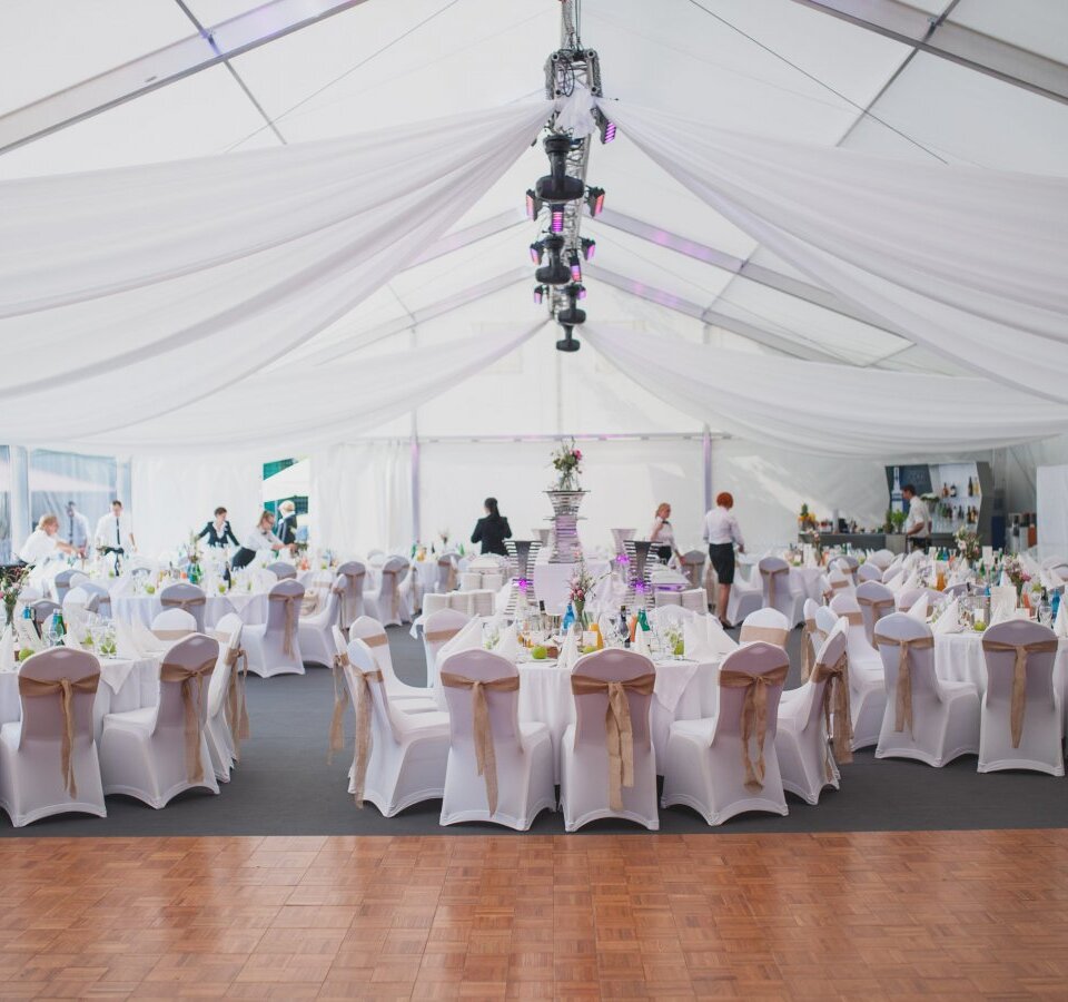 Elegant white marquee with draped ceiling, round tables for a wedding, chairs with beige sashes and floral centerpieces