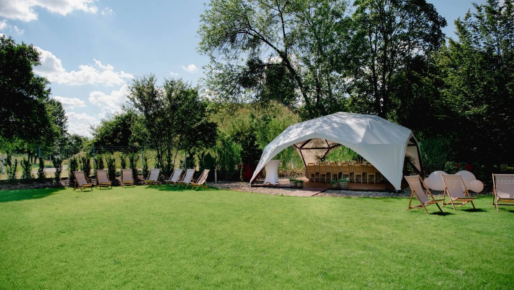 Ogród wokół hotelu z przygotowanym pod dachem stołem dla gości, niedaleko rozstawione leżaki