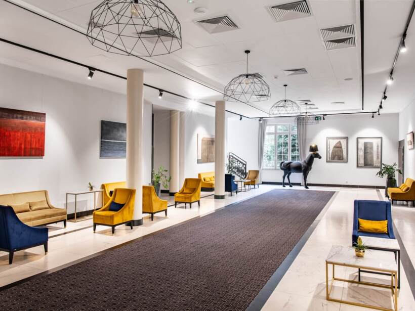 Korytarz hotelowy z jasnym oświetleniem, eleganckimi pomarańczowymi fotelami i rzeźbą