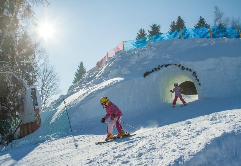 Kinder fahren Ski aus einem in den Schnee gehauenen Tunnel auf einem sonnigen Familienhang mit verschneiten Bäumen