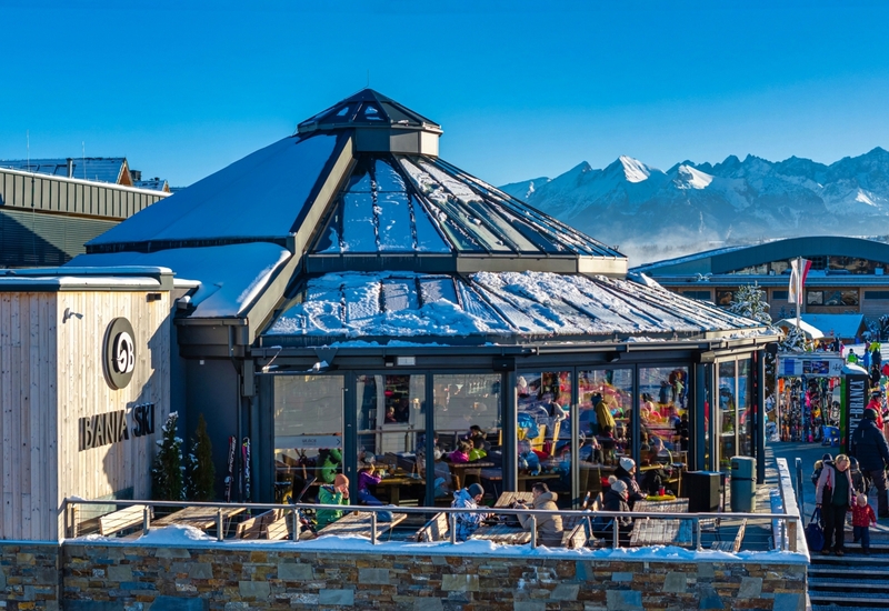 Glasgedecktes Bergrestaurant mit schneebedeckter Terrasse, Gäste speisen an der Piste mit Panorama auf die Gipfel.