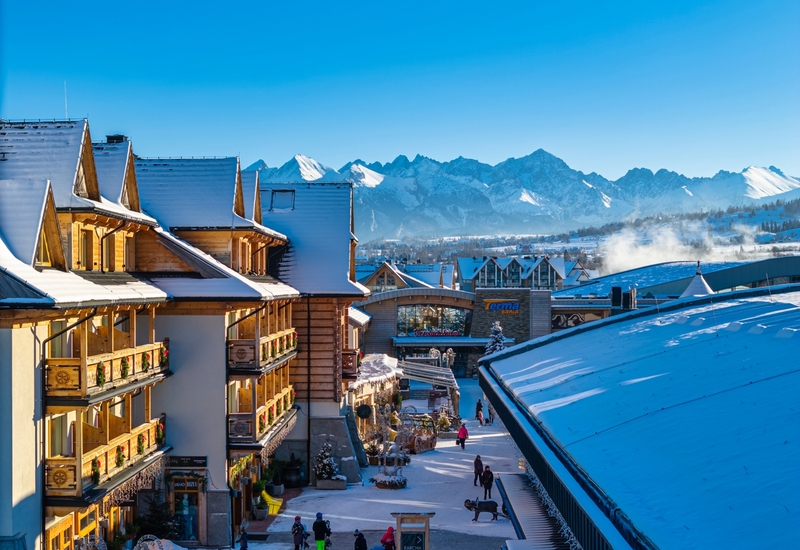 Alpenhotel mit schneebedeckten Dächern und Holz-Balkonen, Blick auf die Hohe Tatra, sonniger Platz mit Geschäften und Eingang zu den Thermen.