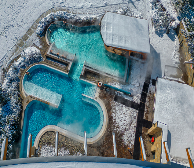 Beheizte Außen-Spa-Pools mit dampfendem türkisfarbenem Wasser, verbundenen Terrassen und einer überdachten Poolbar in einem verschneiten Bergresort