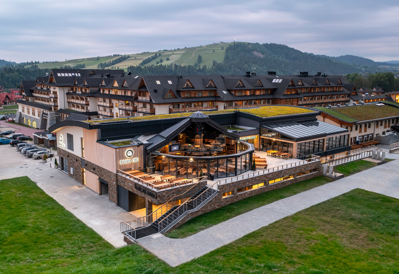 Alpenresort mit verglaster Restaurantterrasse, begrünten Dächern, Zimmerbalkonen und weitem Blick auf die umliegenden Hügel.