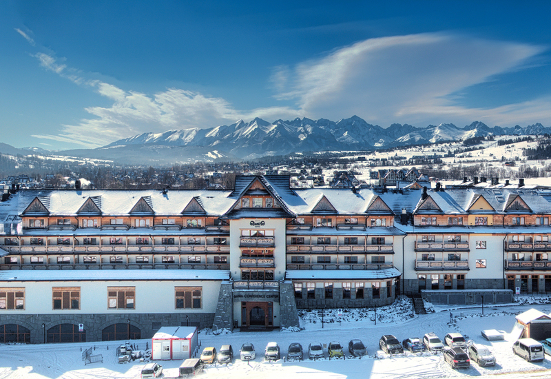 Alpenhotel mit schneebedeckten Dächern und Balkonen, Eingang zum Konferenzzentrum, Parkplatz und Panorama auf verschneite Berge