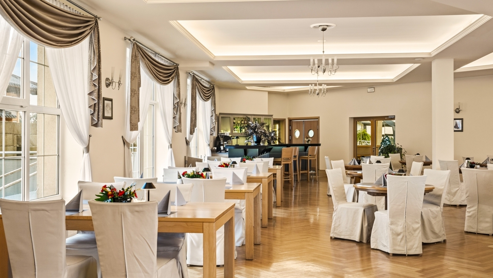 Jasna, elegancka sala restauracyjna z wysokimi oknami w ciężkich zasłonach, drewnianymi stolikami, białymi pokrowcami i barem.