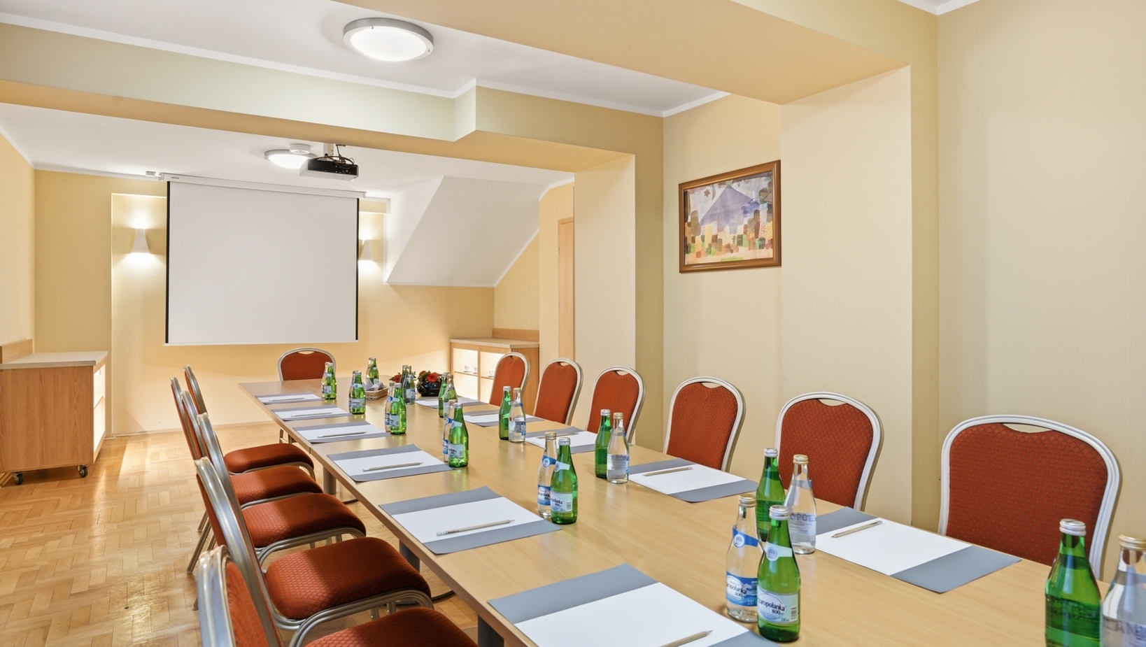 Mała sala konferencyjna z długim stołem, czerwonymi krzesłami, butelkami wody i notatnikami, rzutnik i ekran, ciepłe beżowe ściany