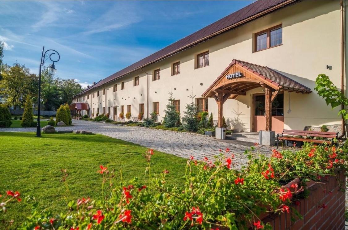 Hotel w stylu wiejskim z drewnianym zadaszonym wejściem, brukowaną ścieżką, zadbaną trawą, czerwonymi kwiatami i ławkami.