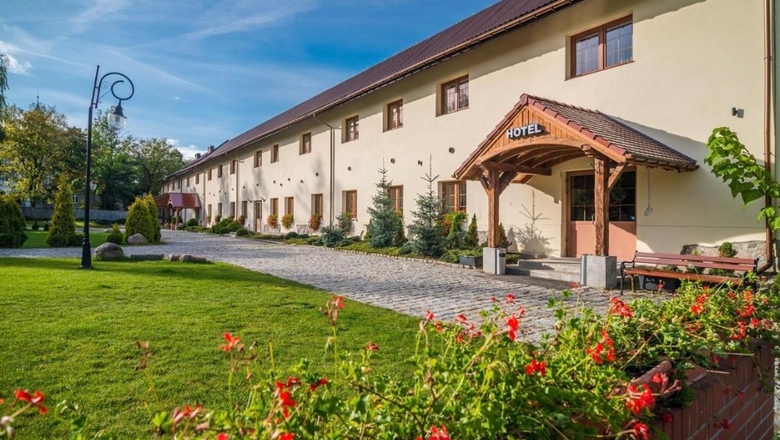 Hotel w stylu wiejskim z drewnianym zadaszonym wejściem, brukowaną ścieżką, zadbaną trawą, czerwonymi kwiatami i ławkami.