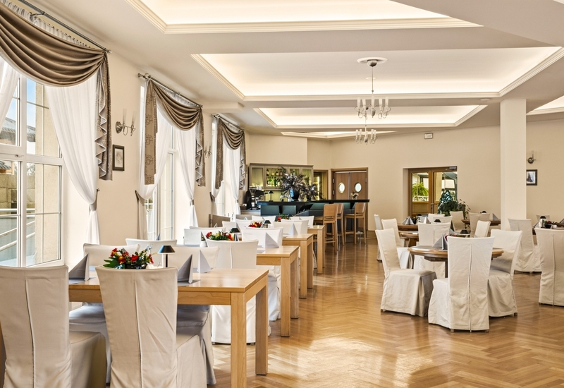 Jasna, elegancka sala restauracyjna z wysokimi oknami w ciężkich zasłonach, drewnianymi stolikami, białymi pokrowcami i barem.