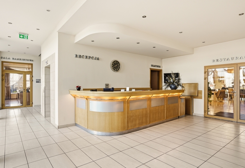 Jasna recepcja z zakrzywioną drewnianą ladą, zegarem, kwiatową dekoracją i przeszklonymi drzwiami do restauracji i sali bankietowej